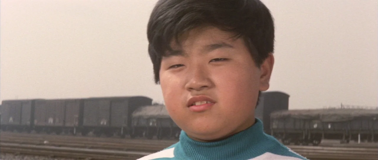 All Monsters Attack (1969) movie still: closeup — A young boy stands in front of a series of cargo train cars on tracks. The background is a hazy,…