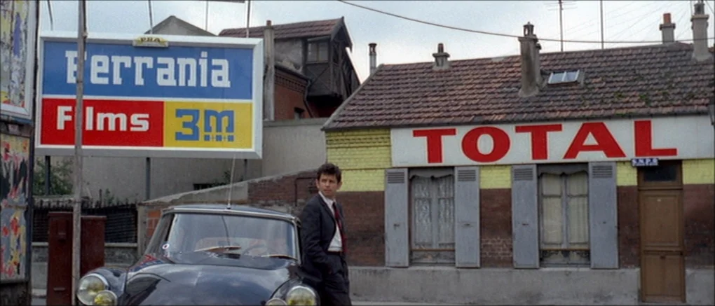 Made in U.S.A. (1966) movie still: medium — A small brick building with a large advertising sign stands behind a dark car. A man in a suit stands…