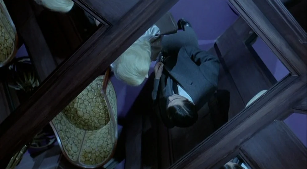 Un Flic (1972) movie still: medium — A narrow corridor with dark wood paneling and purple walls viewed from directly overhead. A blonde woman and a…