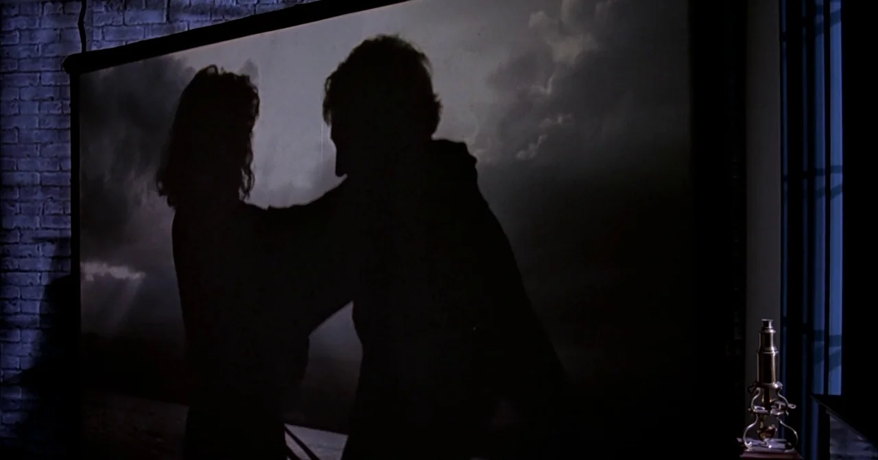 Darkman (1990) movie still: medium — A dark room features a large rear-projection screen displaying a turbulent cloudy sky. A man and woman are…