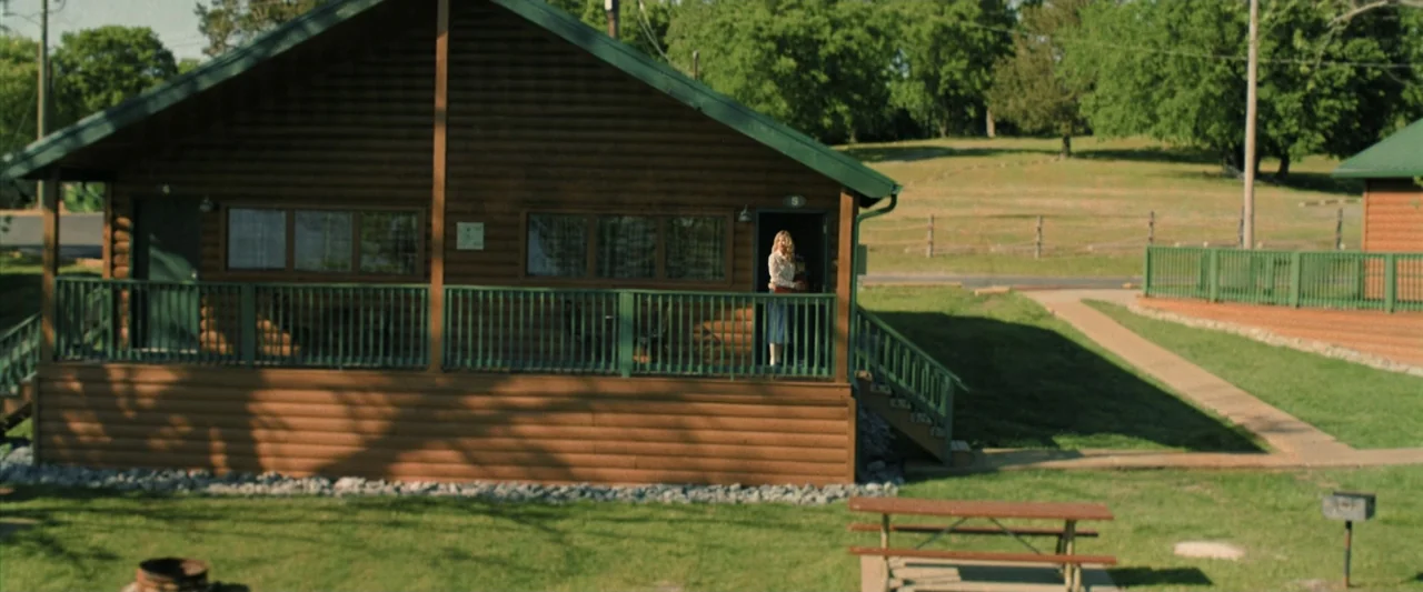 Holland (2025) movie still: wide — A wooden cabin with a green roof stands in a grassy campground. A woman and child stand together in the open…