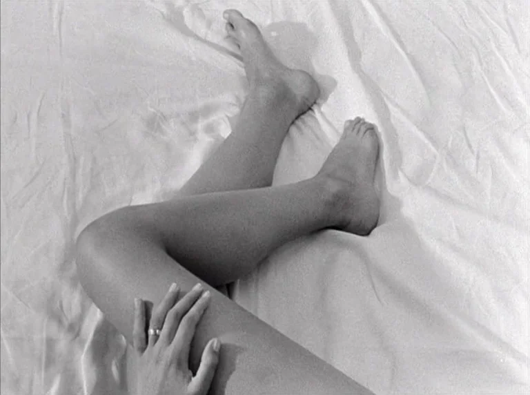 Une Femme Mariée (1964) movie still: closeup — A person's legs rest on a white fabric surface with visible folds. One hand rests on a thigh while…