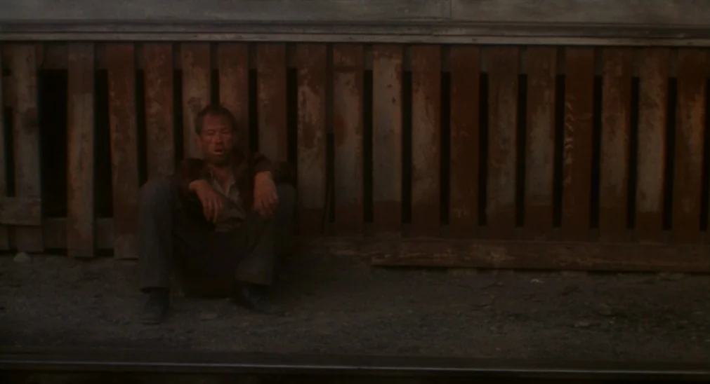 Bound For Glory (1976) movie still: medium — A man sits on the dirt by a weathered vertical wooden plank fence. The area appears to be alongside a…