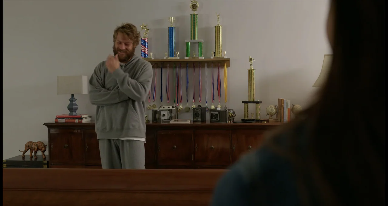 One True Loves (2023) movie still: medium — A living room features a wooden cabinet displaying various sports trophies and ribbons. A bearded man in…