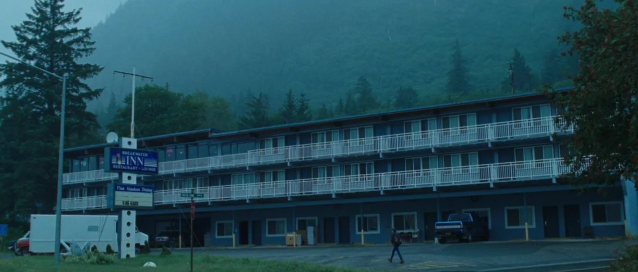 Wildlike (2014) movie still: wide — A long, two-story blue motel building sits at the base of a forested mountain. A single individual walks across…