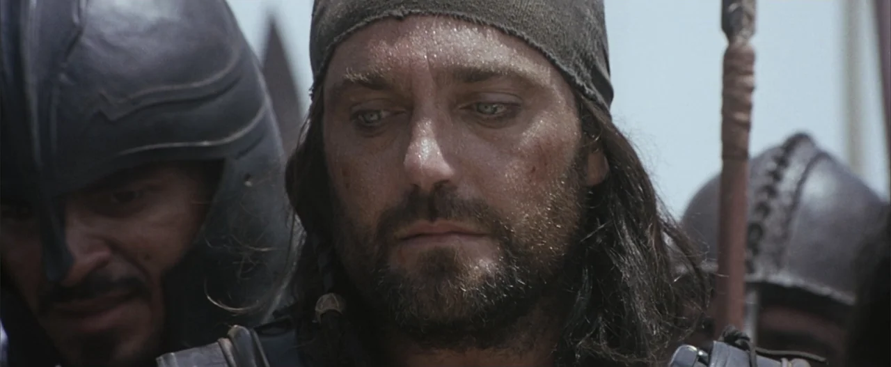 Troy (2004) movie still: closeup — An outdoor battlefield setting with armed men blurred in the background. A bearded man wearing a dark headscarf…