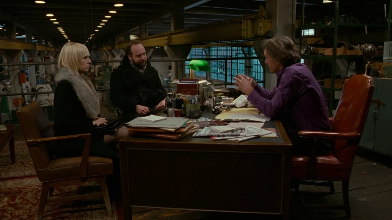 Cold Souls (2009) movie still: wide — A spacious industrial area featuring a desk arrangement positioned in front of factory windows. Three people…