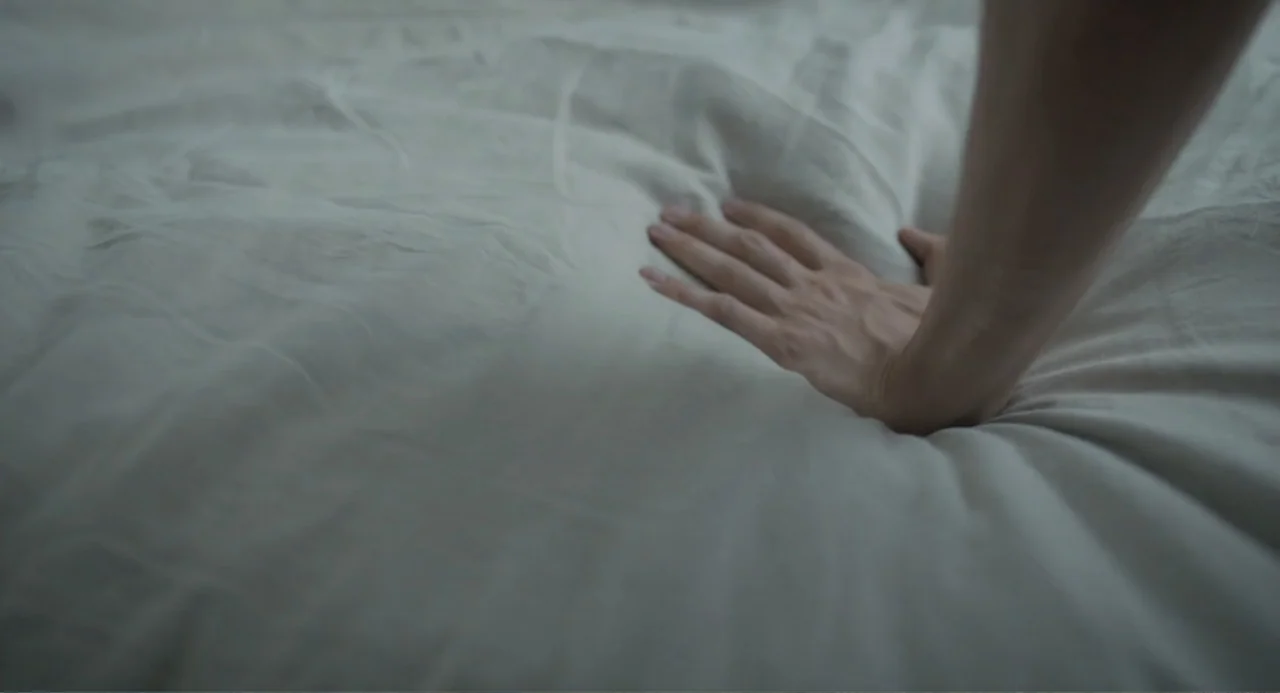 This Closeness (2024) movie still: closeup — A close-up view shows a hand pressing into the soft white fabric of a bed. The wrinkled surface of the…