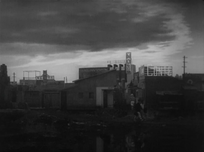Drunken Angel (1948) movie still: wide — A dimly lit street scene features low-slung buildings and prominent rooftop commercial signage. Thick…