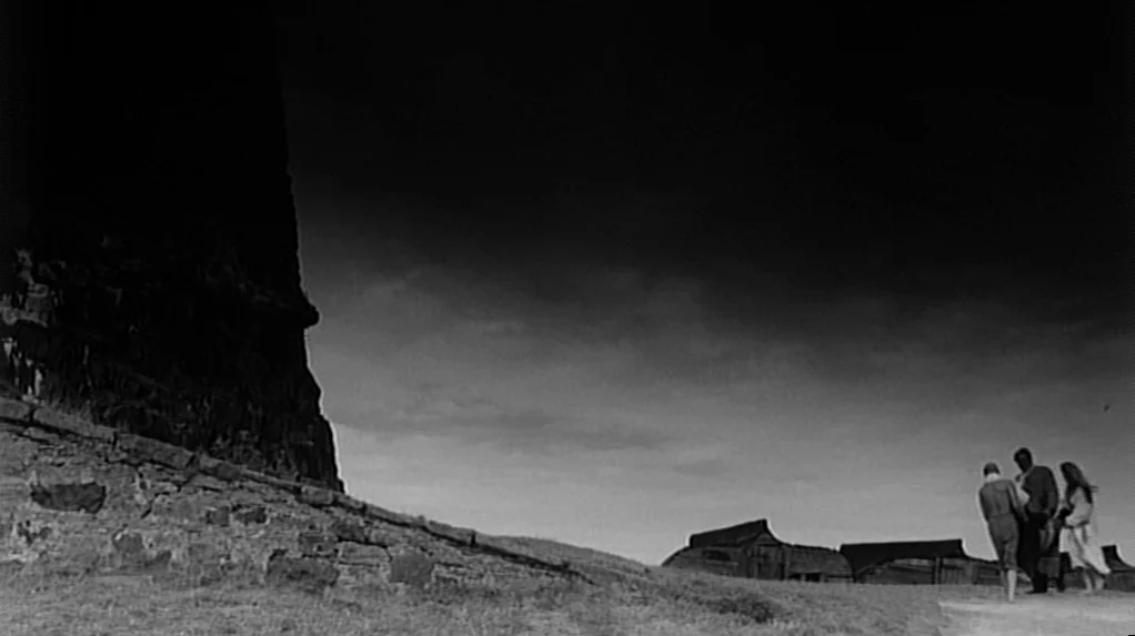 Cul-de-sac (1966) movie still: wide — A dark, imposing rock structure dominates the frame against a stormy sky. Three small figures walk along a…
