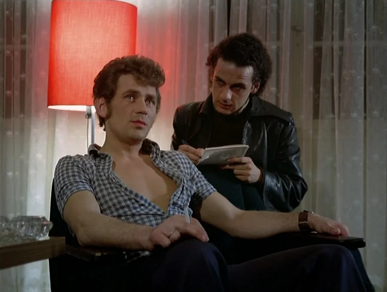 Mother Kusters Goes To Heaven (1975) movie still: medium — A dimly lit room with sheer curtains and a red-shaded lamp behind two men. One man sits…