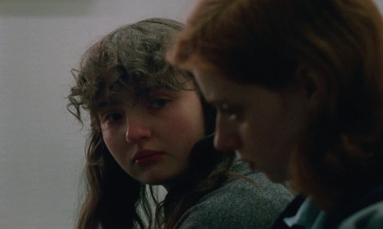 Blue Jean (2022) movie still: closeup — Two women sit side-by-side in a dimly lit, nondescript room. One subject looks directly toward the blurred…