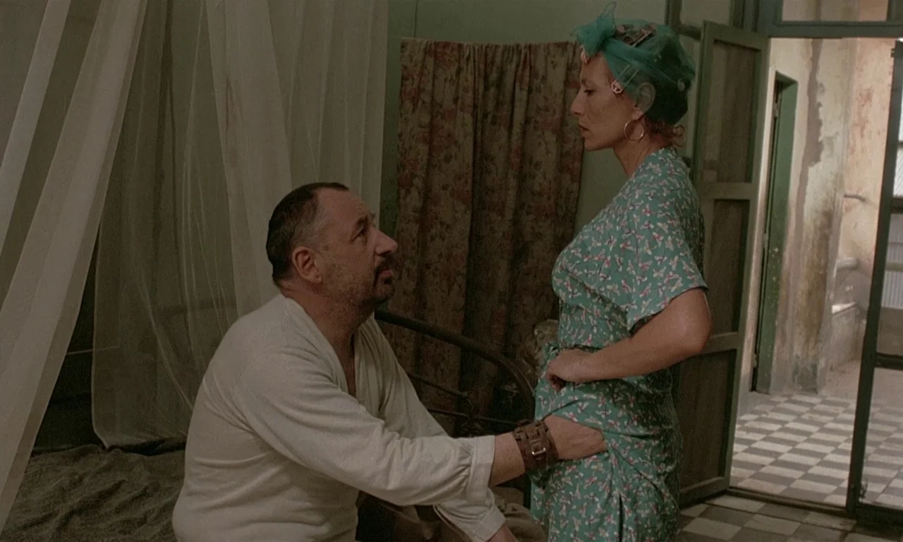 Coup De Torchon (1981) movie still: medium — A sparsely furnished bedroom features a metal bed frame and thin white mosquito netting. A man kneels…