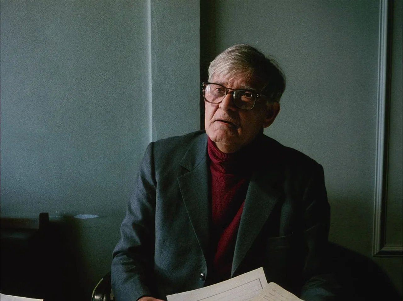 Close-Up (1990) movie still: medium — A man sits at a desk in a room with plain, grey paneled walls. He holds papers while looking off-screen toward…