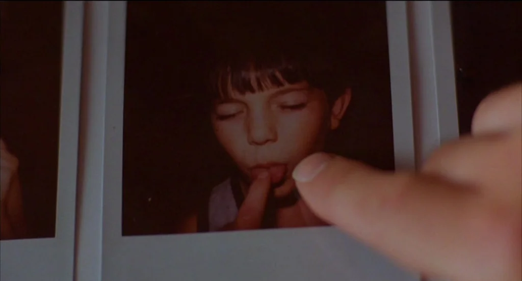 Mysterious Skin (2004) movie still: closeup — A close-up view shows a hand pointing at a physical polaroid photo. The photo depicts a young child…