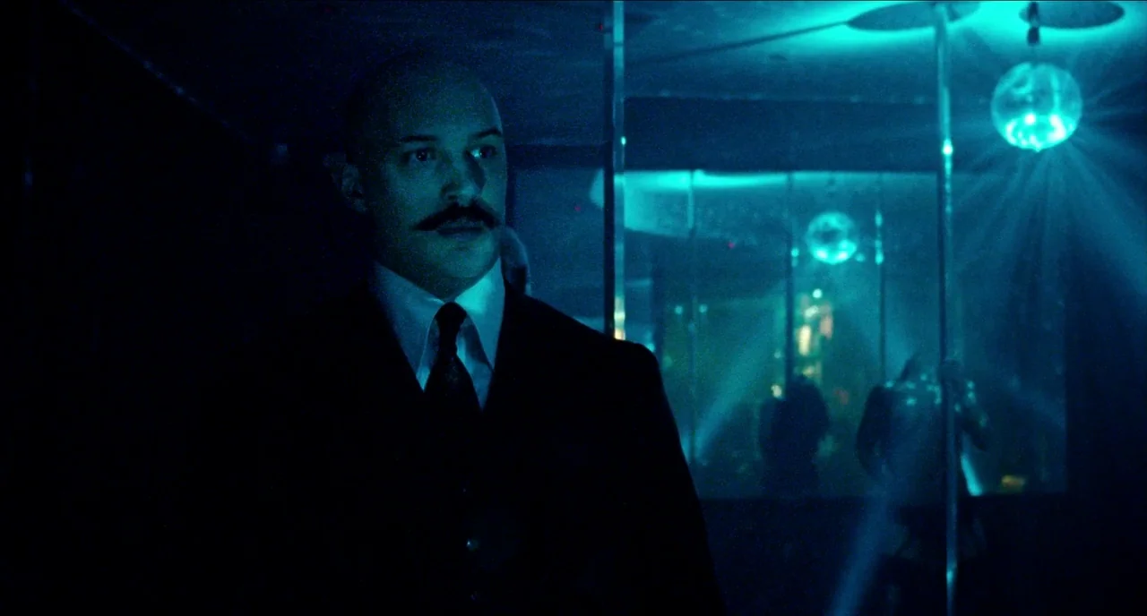 Bronson (2008) movie still: medium — A dark, dimly lit venue featuring mirrored walls and cyan-tinted light fixtures overhead. A man in a dark…