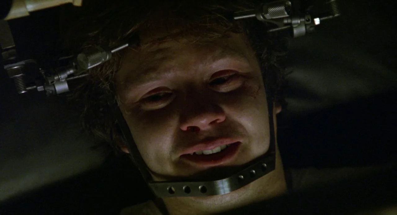 Jacob’s Ladder (1990) movie still: closeup — A dark clinical setting with metallic equipment attached directly to the subject's skull. The man wears…