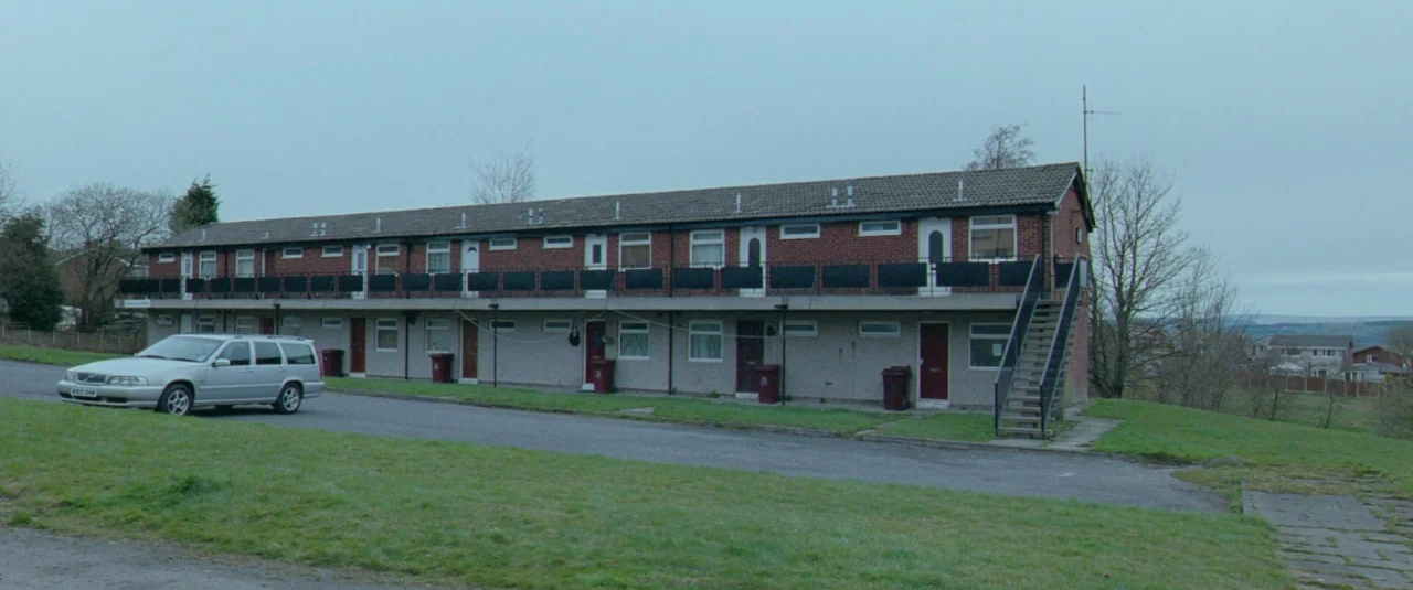 Catch Me Daddy (2014) movie still: wide — A two-story red brick apartment building with a metal exterior staircase sits on grass. A silver station…