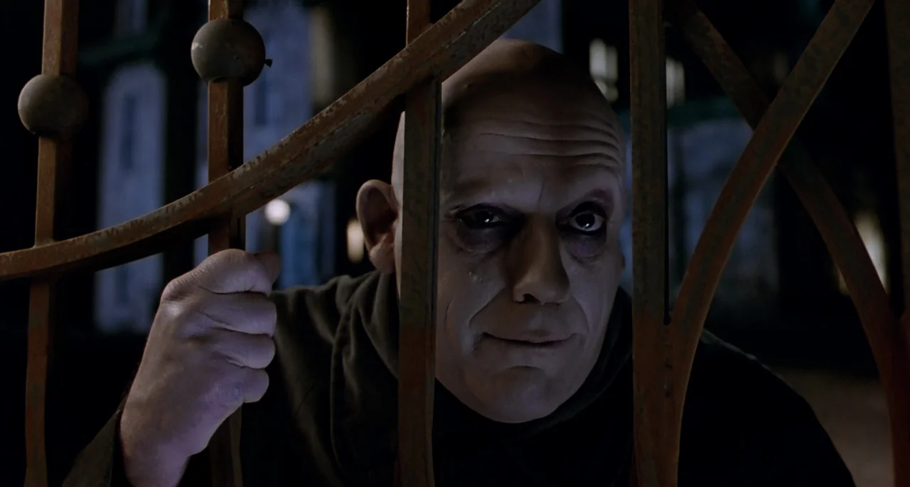 The Addams Family (1991) movie still: closeup — A bald man with heavy eye makeup holds the bars of a rusted gate. The background is a dark,…