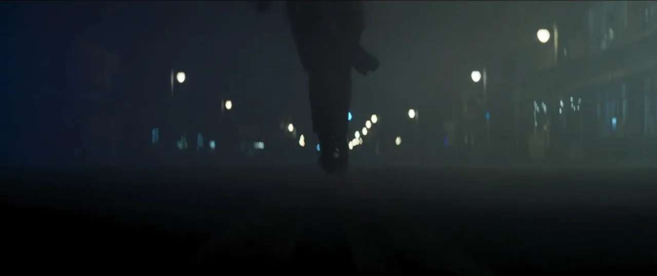 Offseason (2022) movie still: wide — A desolate city street at night is obscured by dense, low-hanging fog. Numerous streetlamps create blurry,…