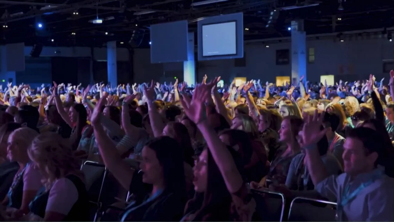 The Rise and Fall of Lularoe (2021) movie still: wide — A dark, expansive indoor event space is filled with a large seated crowd. The audience…