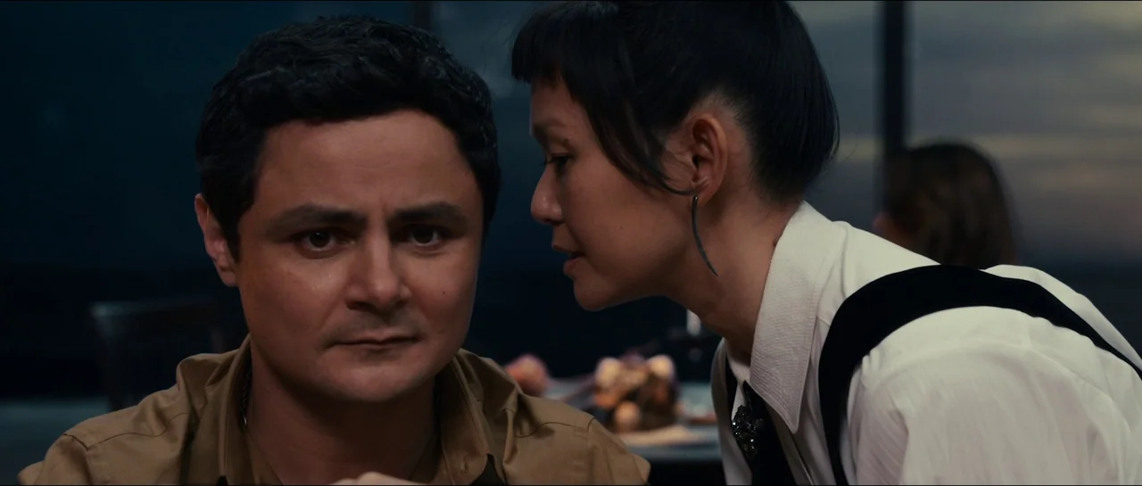 The Menu (2022) movie still: medium — A man and woman sit at a dimly lit dinner table during twilight hours. The woman leans toward the man to speak…