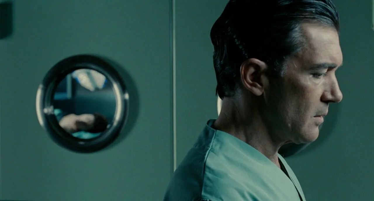 The Skin I Live In (2011) movie still: medium — A man in scrubs stands in front of a flat light green wall. A small circular window shows a…