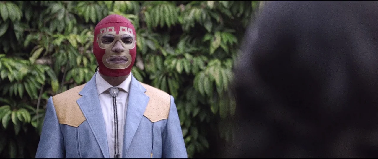 Lowlife (2017) movie still: medium — A man wearing a red and gold luchador mask stands against a background of green leaves. The man in the mask…