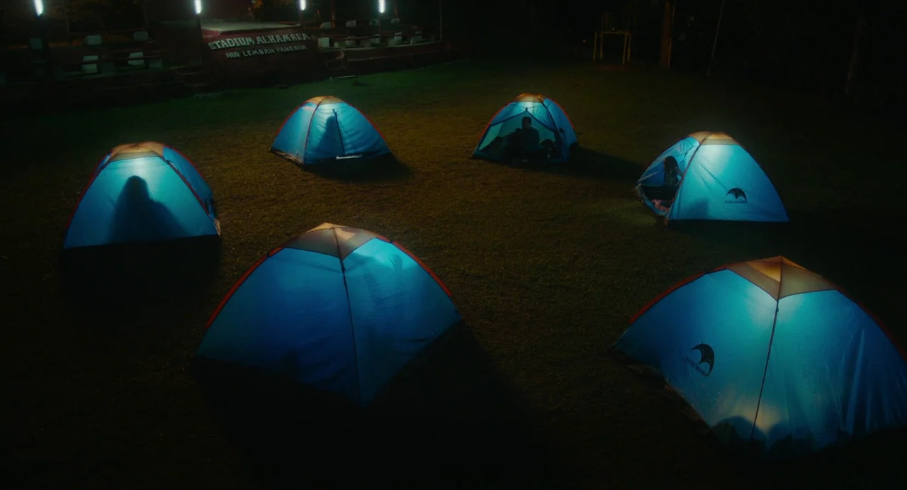 Tiger Stripes (2023) movie still: wide — Six blue camping tents are arranged in a circle on a dark, grassy field. People are visible sitting inside…