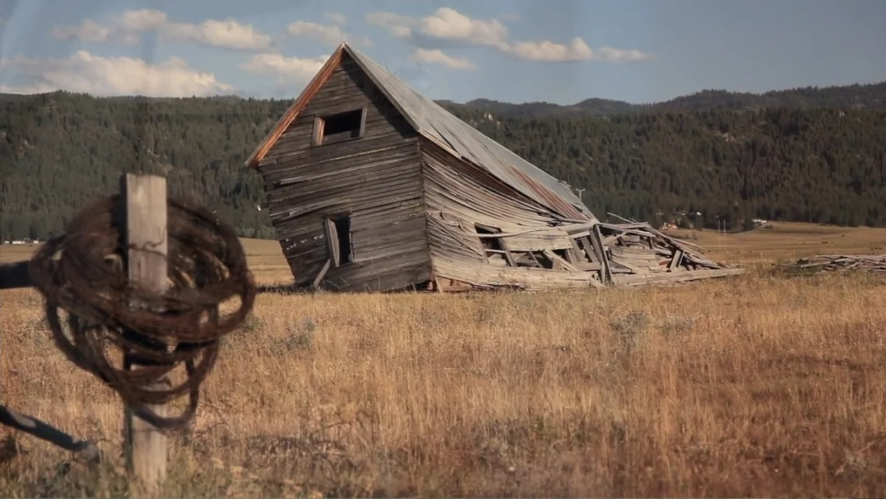 Idaho: The Movie (2012) movie still: wide — A dilapidated wooden barn sits in a vast open field with mountains in background. Barbed wire rests on a…