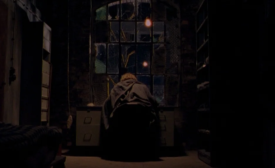 Pola X (1999) movie still: wide — A dimly lit room with brick walls features metal shelving and a central desk. A figure sits at the desk, facing a…