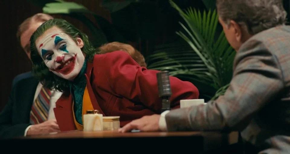 Joker (2019) movie still: medium — A late night television show set with a wooden desk and palm plants. A man in clown makeup smiles while leaning…