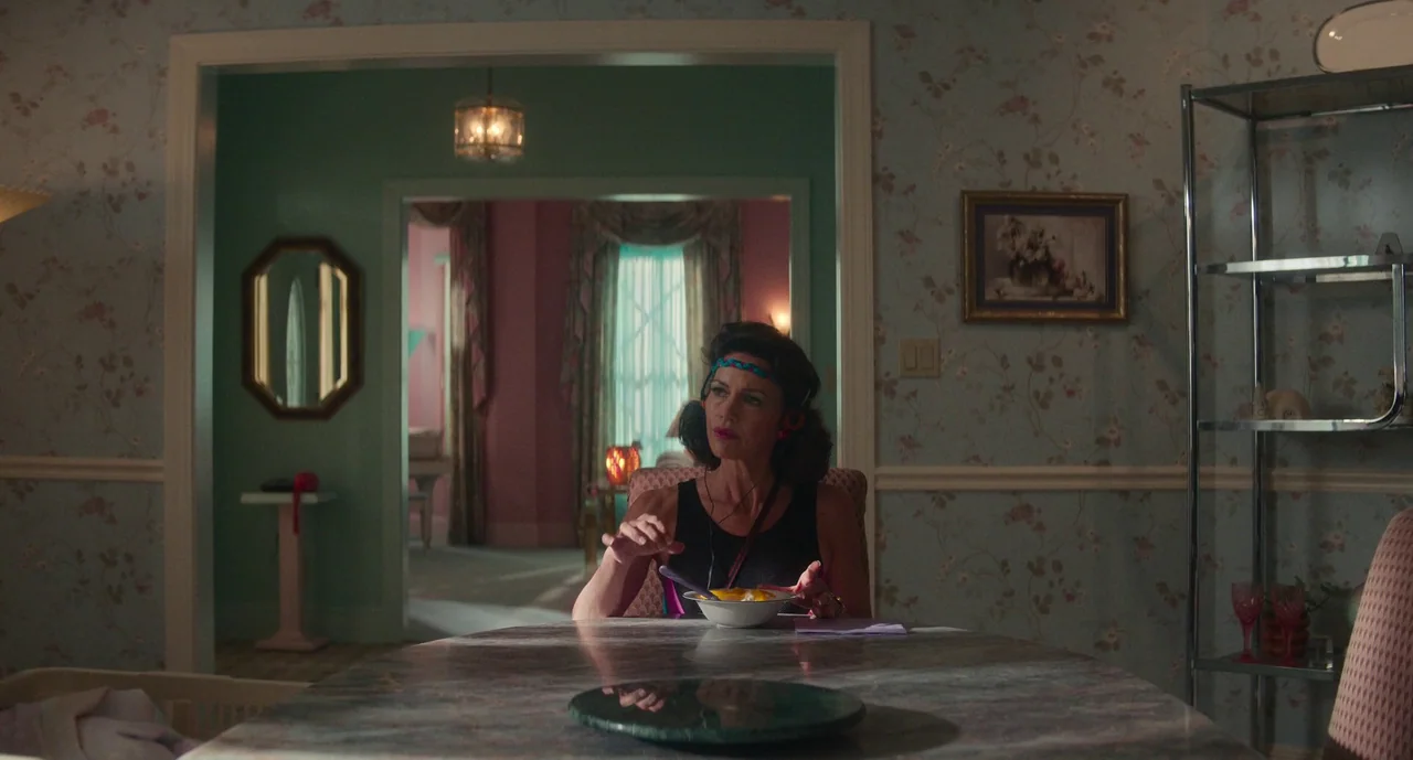 Lisa Frankenstein (2024) movie still: medium — A dining area with pale floral wallpaper and a marbled table centerpiece. A woman sits at the table…