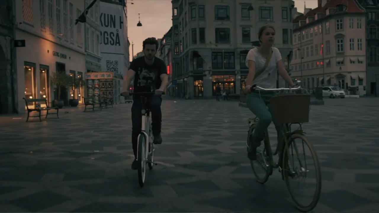 Copenhagen (2002) movie still: fullbody — A European plaza with traditional stone buildings and a patterned stone floor at dusk. A young man and…