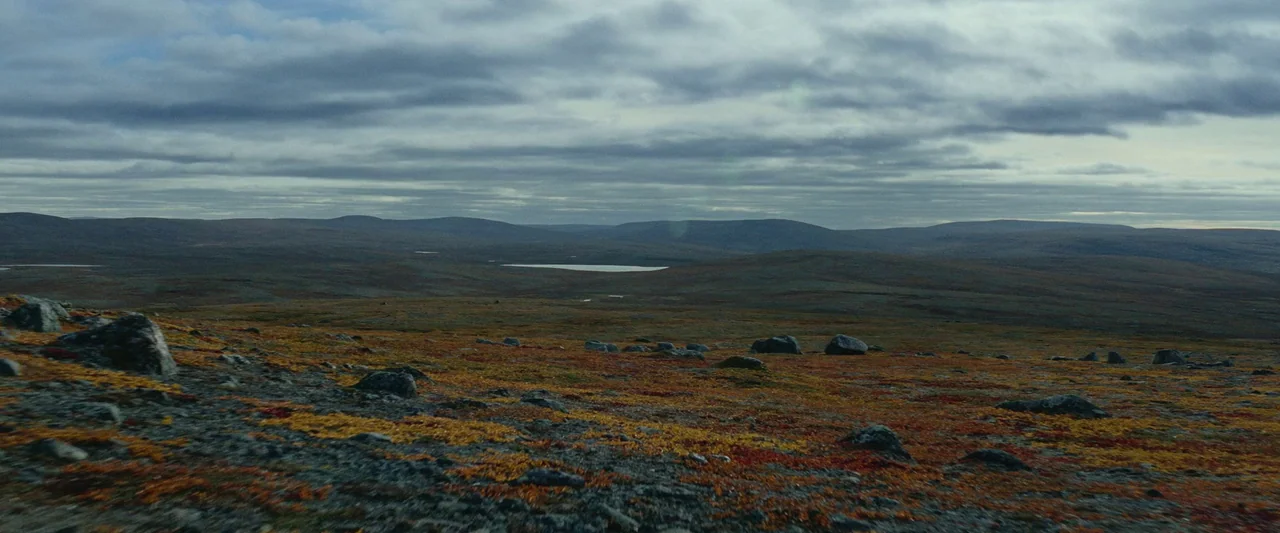 Sisu (2022) movie still: establishing — A vast, rocky tundra under an expansive, heavily clouded sky. The terrain features patches of orange and red…