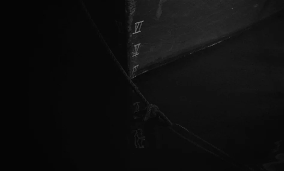 The Man From London (2007) movie still: closeup — A dark, submerged wooden piling with white painted roman numerals sits in deep water. A taut…