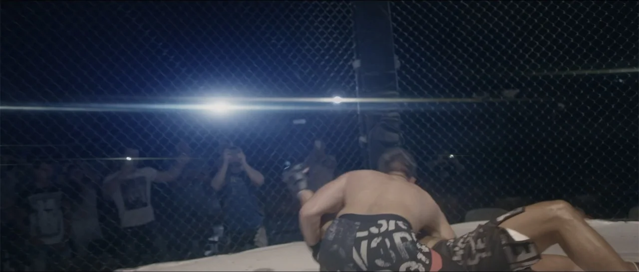 All She Wrote (2018) movie still: medium — An indoor combat ring is enclosed by heavy metal chain-link fencing. Two fighters engage in a grappling…