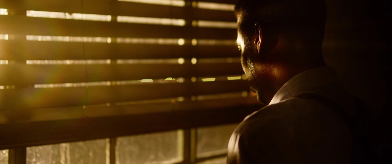 Spiral (2021) movie still: medium — A dimly lit room features a window covered by horizontal venetian blinds. A man stands in profile while looking…