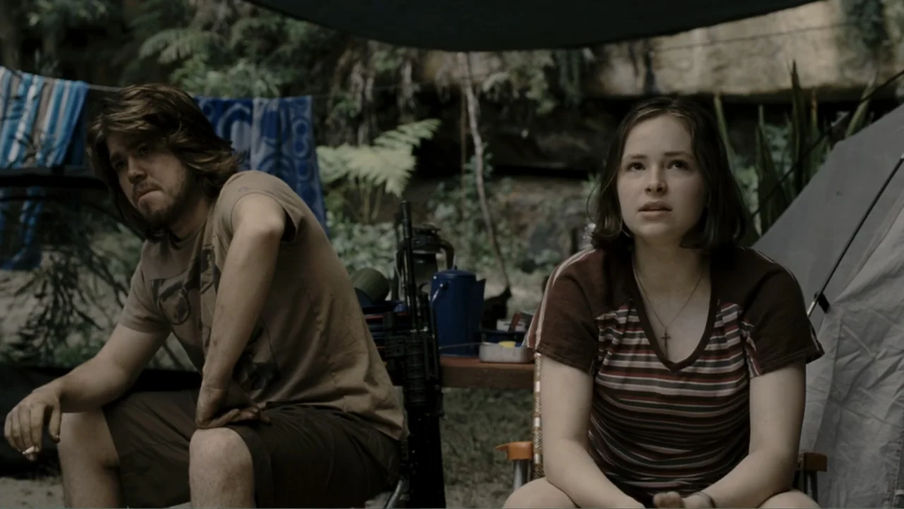 Tomorrow, When the War Began (2010) movie still: medium — A campsite in a rocky, wooded area features a tent and outdoor gear. A young man and woman…