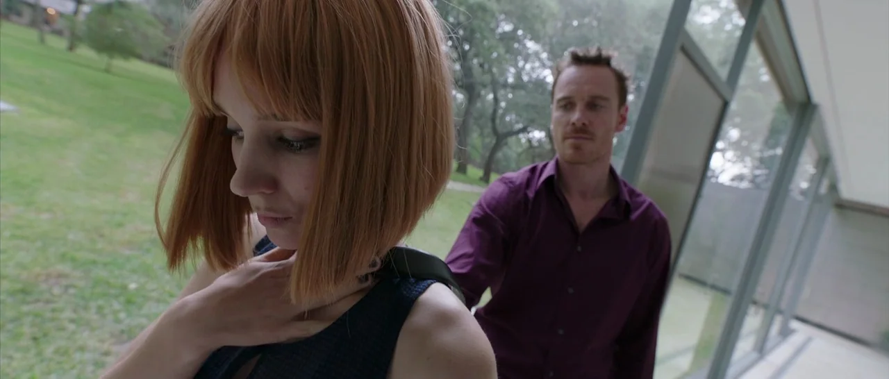 Song to Song (2017) movie still: medium — A modern glass-walled structure faces out onto a green lawn and trees. A red-haired woman foreground…