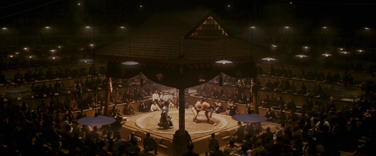 Memoirs of a Geisha (2005) movie still: wide — An indoor sumo arena with a raised clay ring covered by a shinto-style roof. Two heavy-set wrestlers…