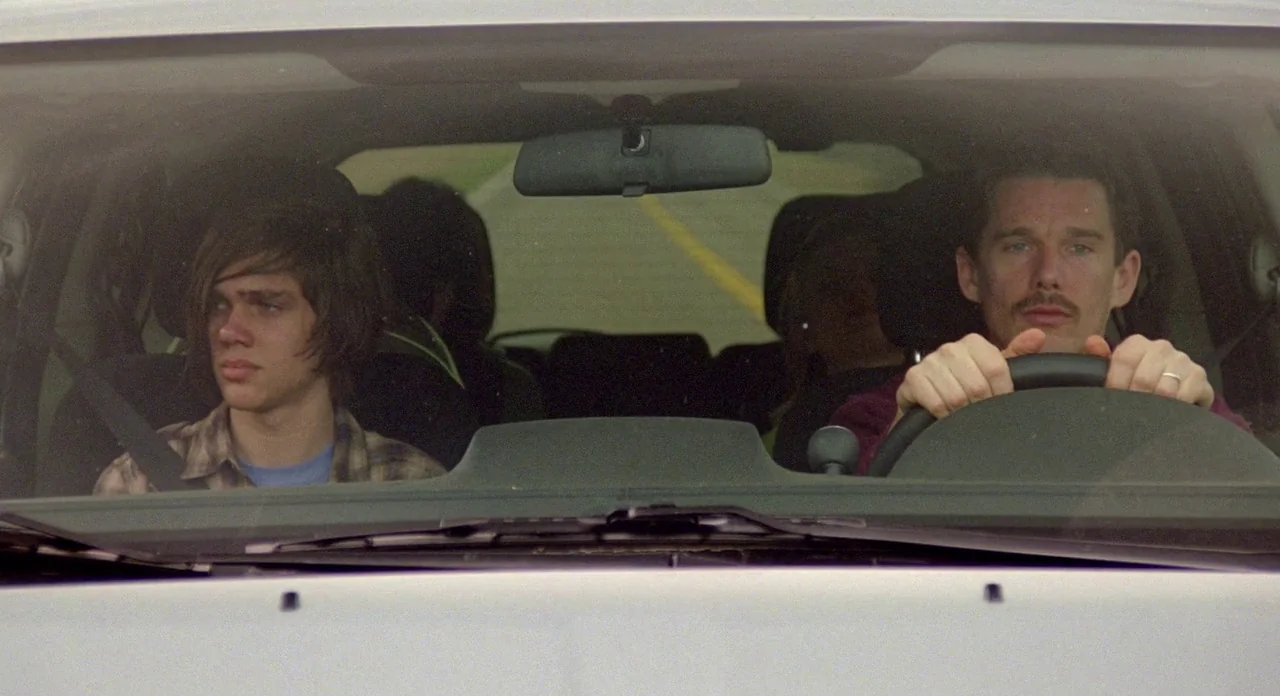 Boyhood (2014) movie still: medium — A view from outside the windshield of a car driving down a road. An older man operates the steering wheel while…