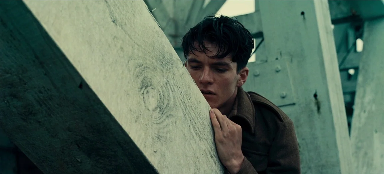Dunkirk (2017) movie still: medium — A young soldier leans against a large, weathered concrete support pillar in an industrial area. He rests his…