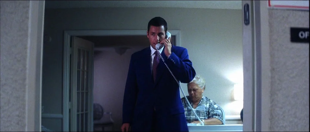 Punch Drunk Love (2002) movie still: medium — An interior shot looking through a doorway into a muted lobby space. A man stands in the foreground on…