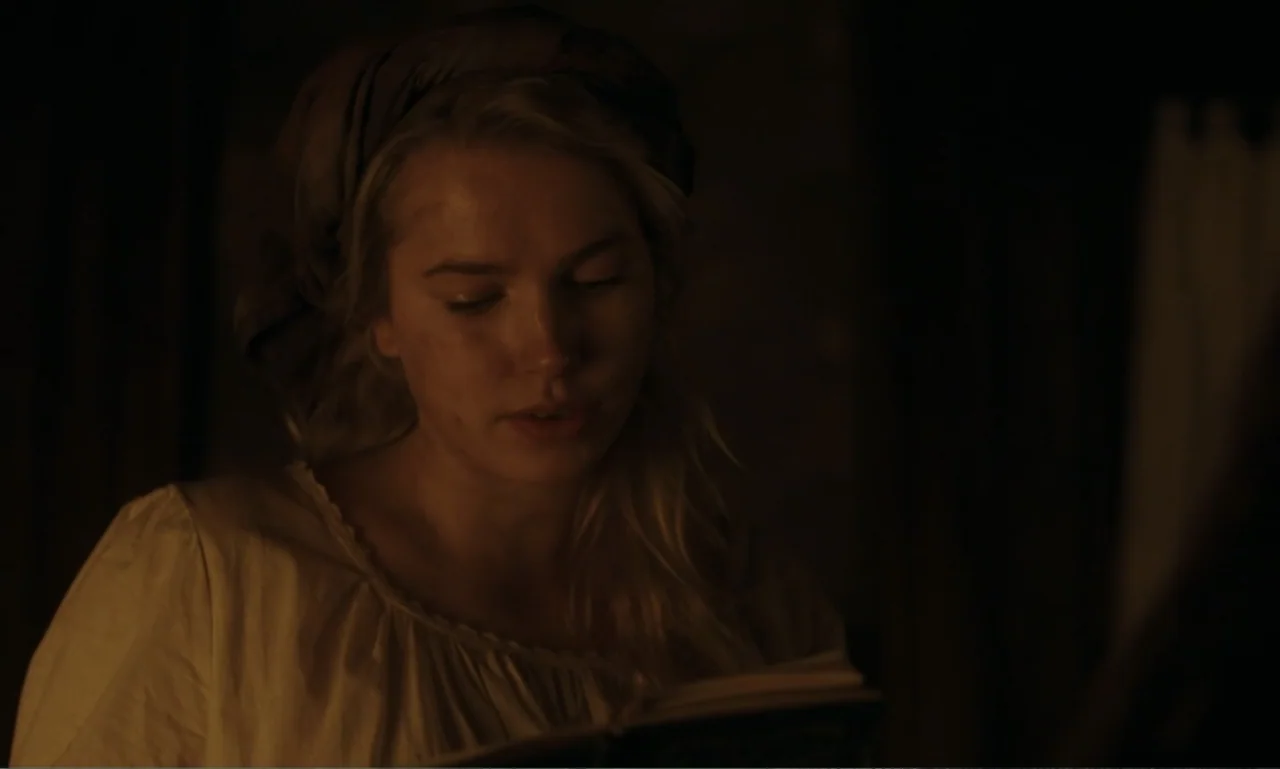 The Ugly Stepsister (2025) movie still: closeup — A dimly lit interior room with shadows obscuring the surroundings. A young woman wears a bonnet…