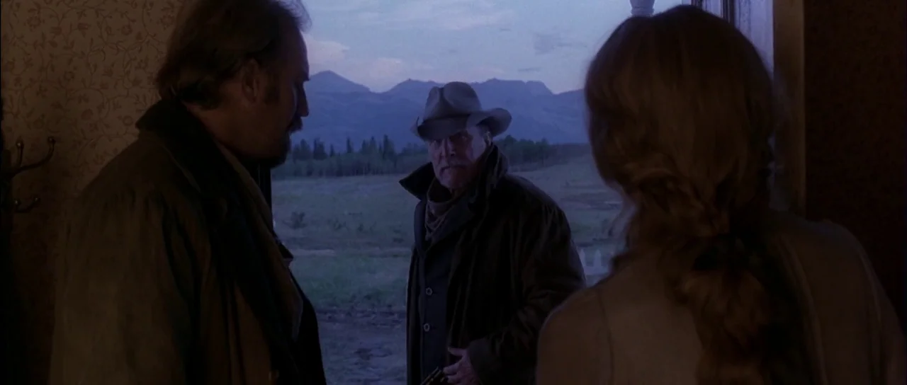 Open Range (2003) movie still: medium — A man in a cowboy hat stands outside a dark doorway at dusk. Mountain peaks are visible in the background…