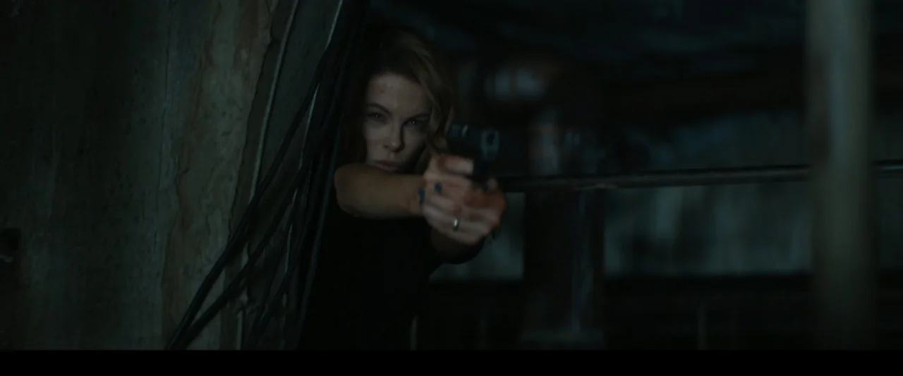Canary Black (2024) movie still: medium — A dark, dimly lit basement with large metal pipes and industrial wiring visible. A woman holds a handgun…