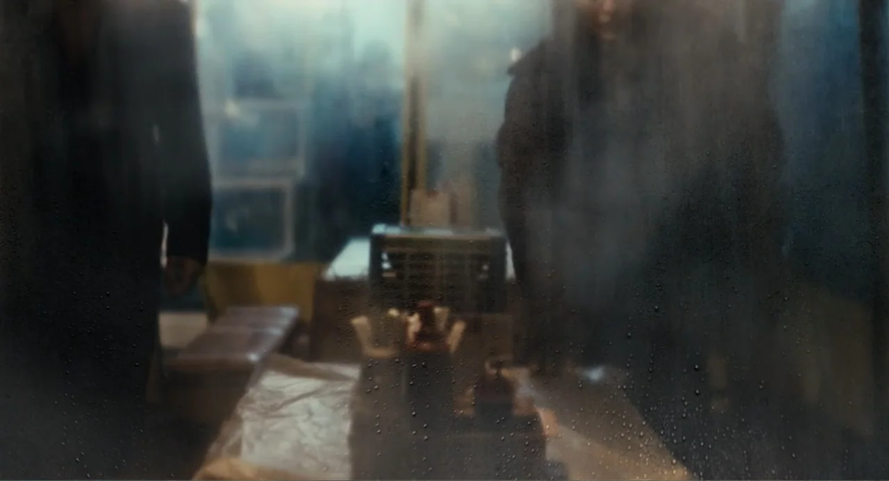 Winter in Sokcho (2025) movie still: medium — A rain-streaked window obscures the interior of a dark, dimly lit diner. Two figures in dark coats…