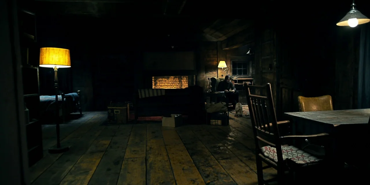 Polar (2019) movie still: wide — A dark, rustic cabin interior features wood floors, a brick fireplace, and scattered furniture. A lone person sits…