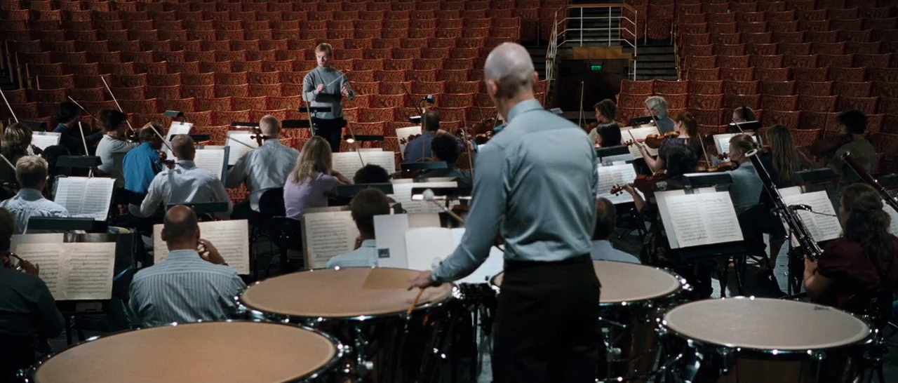 Sound of Noise (2010) movie still: wide — An orchestra sits on a large stage in a concert hall with red seating. A man in the foreground plays the…